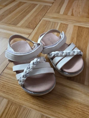 Vita sandaler med flätad rem - Snygga vita sandaler med flätad rem i vitt och silver. Sandalerna har flera remmar över foten och justerbar kardborrestängning. Perfekta för sommaren!