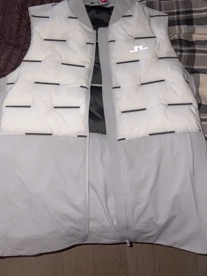 Ljusgrå dunväst från J.Lindeberg - Snygg ljusgrå dunväst från J.Lindeberg med vita partier och svarta horisontella detaljer. Västen har en hög krage, dragkedja framtill och en modern, boxig passform. Perfekt för lager-på-lager och streetwear-vibe.