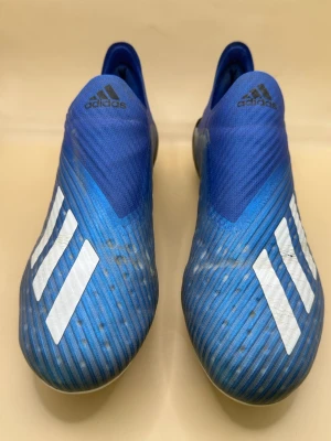 Adidas X 19+ FG  - Säljer ett par blå Adidas X fotbollsskor med vita detaljer och klassiska tre ränder på sidan. Skorna har en strömlinjeformad design utan snörning och är tillverkade i syntetmaterial för bästa passform och grepp på planen. Perfekta för dig som vill ha speed och kontroll.