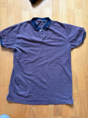 Lila/grey pikétröja från Tommy Hilfiger - Säljer en lila pikétröja från Tommy Hilfiger med klassisk krage och knappar framtill. Tröjan har korta ärmar och den ikoniska lilla loggan broderad på bröstet. Materialet känns som bomull och kragen är mörkblå för en snygg kontrast.(är mer som L/XL)