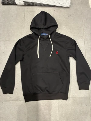 Svart hoodie från Polo Ralph Lauren - Svart hoodie från Polo Ralph Lauren med vit dragsko och klassisk röd broderad logga på bröstet. Hoodien har känguruficka framtill och ribbade muddar vid ärmslut och nederkant. Perfekt för en chill och stilren look.