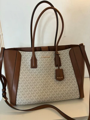 Michael Kors brun och beige handväska - Stilren handväska från Michael Kors i beige canvas med MK-logga över hela väskan och bruna detaljer i läder. Väskan har två långa handtag, axelrem och flera innerfack. Perfekt för dig som vill ha både snygg och praktisk väska till vardagen.
