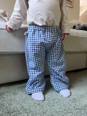 Rutiga blåvita mjukisbyxor - Säljer ett par mjukisbyxor med blåvitt rutigt mönster och resår i midjan. Byxorna har en avslappnad passform och är tillverkade i ett mjukt, bekvämt tyg som passar perfekt för chill dagar hemma. Perfekt för dig som gillar retrovibbar och sköna plagg.