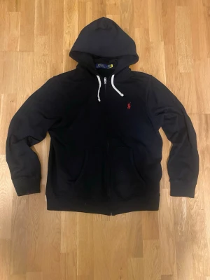 Svart hoodie från Polo Ralph Lauren - Klassisk svart hoodie från Polo Ralph Lauren med dragkedja, vit dragsko i huvan och den ikoniska röda logobrodyr på bröstet. Hoodien har kängurufickor och är tillverkad i mjukt bomullsmaterial. Perfekt för en avslappnad och stilren look.
