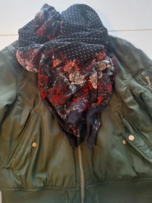 Svart sjal med prickar och rosor - Säljer en stor, fyrkantig sjal i svart med vita prickar och ett färgstarkt mönster av röda och vita rosor längs kanterna. Materialet känns mjukt och lätt, perfekt att svepa runt halsen för en cool och personlig stil. Ca 110x110 cm