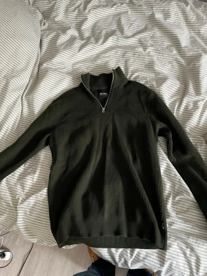 Halfzip  - Snygg mörkgrön ribbad tröja  med halv dragkedja och hög krage. Perfekt för lager på lager och enkel att matcha med jeans eller chinos. Tröjan har långa ärmar och en stilren look.