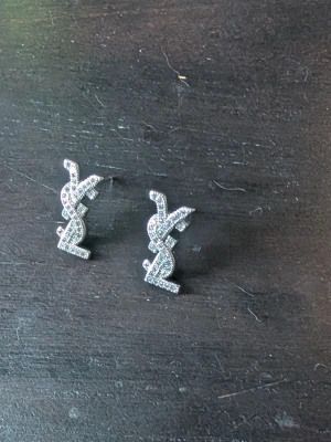 Örhängen med strass - Säljer ett par snygga örhängen i silverfärg med glittrande strass. Örhängena har den ikoniska YSL-loggan och är perfekta för dig som gillar statement-smycken. Materialet är metall med små gnistrande stenar som ger extra lyxig känsla.