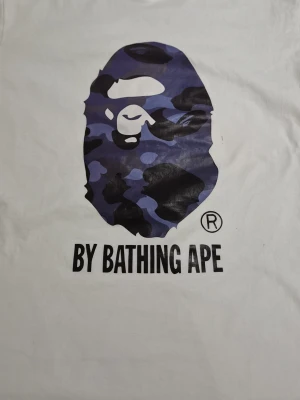 Vit BAPE t-shirt med blått camomönster - Säljer en vit t-shirt från By Bathing Ape med det ikoniska apansiktet i blått och svart camomönster på bröstet. T-shirten har klassisk passform och korta ärmar. Perfekt för dig som gillar streetwear och vill sticka ut.