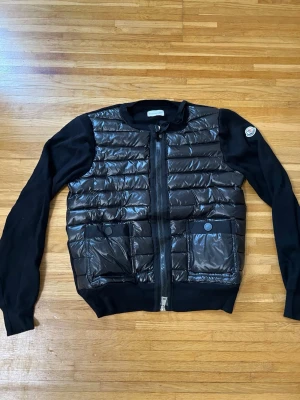 Svart dam-cardigan från Moncler - Snygg svart dunväst från Moncler med quiltad front, två stora fickor med tryckknappar och dragkedja framtill. Västen har stickade svarta ärmar och Moncler-logga på ena ärmen. Materialet är en mix av glansigt dun och mjukt tyg. Perfekt för dig som vill ha en trendig och sportig look. kvitto finns!!