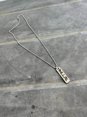 Silverfärgat halsband med Billie-platta - Snyggt halsband i silverfärg med en rektangulär platta där det står 'BILLIE' i svart graffiti-liknande text. Kedjan består av små kulor och ger ett modernt streetwear-inspirerat intryck. Perfekt accessoar för dig som gillar personlig stil.
