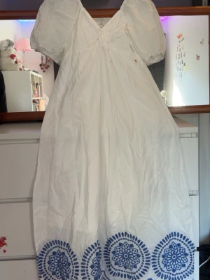 Vit långklänning med blå broderi - Vit långklänning med puffärmar och v-ringad front. Klänningen har blå broderade mönster längs nederkanten och en lätt volangform. Perfekt för sommarens alla tillfällen, med en luftig och romantisk look.