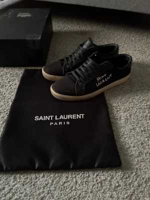 Svarta Saint Laurent sneakers - Snygga svarta sneakers från Saint Laurent med vit logga på sidan och beige sula. Klassisk låg modell med svarta skosnören och stilren design. Kommer med originalpåse och låda. Perfekta för dig som gillar exklusiva och tidlösa skor. Storlek 43! Pris kan diskuteras