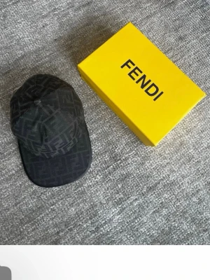 Svart Fendi keps med FF-mönster - Snygg svart keps från Fendi med det klassiska FF-logomönstret över hela kepsen. Skärmen har en stilren svart kant i läderimitation. Perfekt accessoar för dig som gillar lyxiga detaljer och streetstyle.