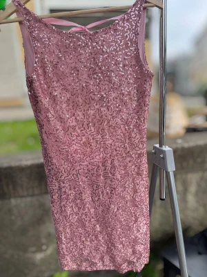 Rosa paljettklänning med öppen rygg - Elegant rosa klänning med vackra paljettdetaljer över hela plagget. Klänningen har en djup, öppen rygg med ett dekorativt satinband som knyts upptill och ger en feminin form. Perfekt till fest, nyår, bröllop eller andra speciella tillfällen.