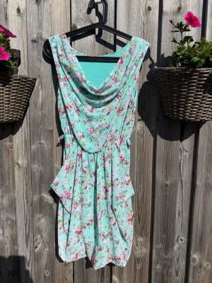 Blommig mintgrön kortklänning (S) - Söt mintgrön kortklänning med blommigt mönster i rosa och vitt. Klänningen har draperad urringning, knyt i midjan och volangdetaljer på sidorna. Perfekt för varma dagar och ger en romantisk vibe.