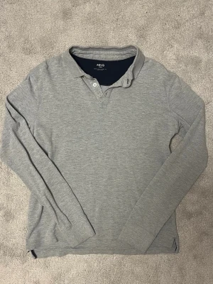 Grå långärmad tröja från MNG MAN - Grå långärmad tröja från MNG MAN med klassisk krage och knappar vid halsen. Tröjan har en enkel och stilren design, perfekt för en avslappnad look. Tillverkad i mjukt bomullsmaterial för extra komfort.