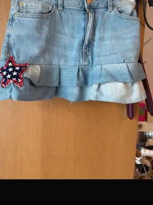 Jeanskjol med volang och stjärndetalj - Söt ljusblå jeanskjol med volangkant och råa kanter. Pris kan diskuteras!💖