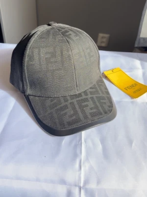 Grå keps från Fendi med FF-mönster - Snygg grå keps från Fendi med det klassiska FF-logomönstret över hela kepsen. Kepsen har böjd skärm och justerbar baksida. Tillverkad i ett slitstarkt tyg som ger en exklusiv känsla. Perfekt för dig som vill ha en stilren accessoar med lyxig touch.