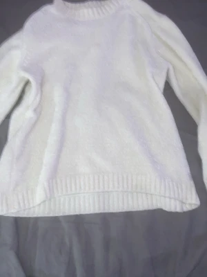 Vit stickad tröja med ribbade detaljer - En enkel och stilren vit stickad tröja med rund hals och ribbade muddar vid ärmslut och nederkant. Tröjan har lång ärm och är gjord i ett mjukt material som känns skönt mot huden. Perfekt för lager-på-lager och att hålla sig varm under kyliga dagar.