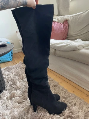 Svarta overknee stövlar med klack - Säljer ett par svarta overknee stövlar med hög klack och rundad tå. Stövlarna är i ett mjukt mockaliknande material och har en stilren, slimmad passform som går högt upp över knät. Perfekta för dig som vill ha ett statement-plagg till din outfit.