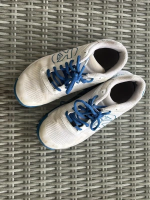 Vita sneakers från Kempa  - Säljer ett par vita träningsskor från Kempa med blå snören och blå sula.  Perfekta för träning eller sportaktiviteter.