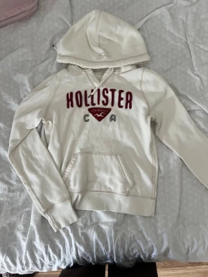 Vit hoodie från Hollister med tryck - Snygg vit hoodie från Hollister med röd broderad logga och text på bröstet. Klassisk känguruficka framtill och huva med dragsko. Tillverkad i mjukt bomullsmaterial, perfekt för chill dagar eller när du vill ha en avslappnad look.