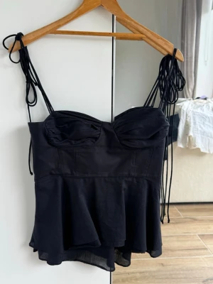 Svart linnetopp med knytband - Snygg svart linnetopp med tunna axelband som knyts på axlarna. Toppen har en figurnära passform upptill, markerade kupor och en volang nedtill som ger en cool och trendig look. Perfekt att styla med jeans eller kjol.