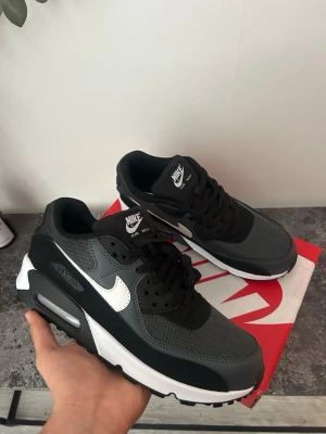 Nike Air Max 90 svart/grå sneaker - Nike Air Max 90 sneakers i svart och mörkgrå mesh och syntetmaterial med vit sula och vit swoosh. Klassisk chunky design med synlig Air-enhet i hälen och svarta skosnören. Perfekt för dig som gillar streetwear och sportig stil.