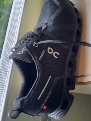 Svarta On Running sneakers - Säljer ett par svarta On Running sneakers med coola detaljer och logga på sidan. Skorna har en unik sula med hål, reflekterande detaljer och texten 'Swiss Engineering'. Tillverkade i slitstarkt textilmaterial och har snörning framtill.