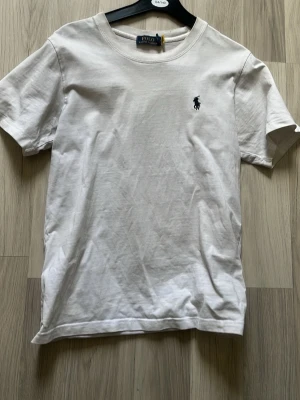 Vit t-shirt från Polo Ralph Lauren - Klassisk vit t-shirt från Polo Ralph Lauren med rund halsringning och den ikoniska broderade loggan på bröstet. Tillverkad i mjuk bomull och har en normal passform med korta ärmar. Perfekt basplagg för en clean och stilren look.