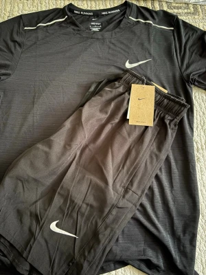 Svart Nike löparset t-shirt & shorts - Nike löparset med svart t-shirt och matchande shorts. Båda plaggen har Nike-logga i vitt och är tillverkade i lätt, snabbtorkande Dri-FIT-material som andas. T-shirten har rund hals och kort ärm, shortsen har resår i midjan och fickor.
