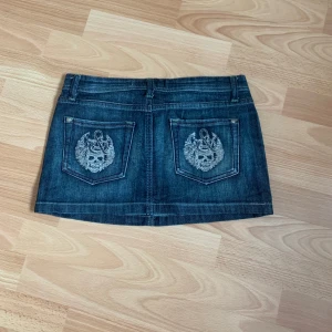 Mörkblå jeanskjol från Top 7 - Kort mörkblå jeanskjol med klassiska fickor fram och bak. Snygga broderade detaljer på bakfickorna och knappstängning framtill. Perfekt för en avslappnad och trendig stil. Längd 26 💗 midja tvärs över 35 💗