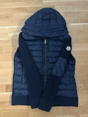 Moncler Cardigan - Snygg mörkblå pufferjacka från Moncler med quiltad framsida, stickade ärmar och rygg samt stor huva. Jackan har dragkedja framtill, två sidofickor och Moncler-logga på ärmen. Materialmix av dun och stickat ger både värme och stil.