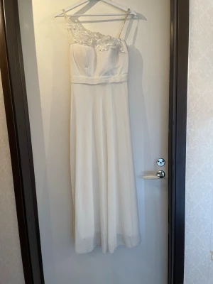 Vit långklänning med spetsdetaljer  - Elegant och lätt vit långklänning med tunna axelband. Överdelen har vackra spetsapplikationer med blommönster och små pärlor. Kjolen faller rakt och mjukt ner till golvet. Perfekt för speciella tillfällen. Har en inbyggd bh vaddering.  