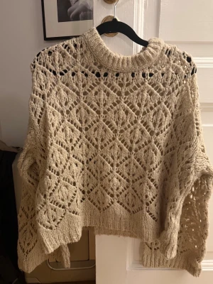 Beige virkad stickad tröja - Trendig beige stickad tröja med virkat mönster och bred passform. Tröjan har rund halsringning och långa ärmar. Perfekt för lager på lager och ger en avslappnad men stilsäker look. Materialet känns mjukt och luftigt.