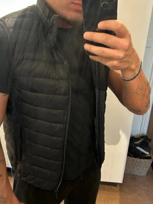 Svart lätt dunväst med dragkedja - Snygg svart dunväst med quiltad design och hög krage. Västen har dragkedja framtill och är tunn men värmande, perfekt att slänga över en hoodie eller t-shirt. Passar dig som gillar en clean och stilren look.
