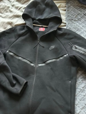 Svart Nike hoodie med dragkedja - Svart hoodie från Nike med hel dragkedja och reflekterande detaljer över bröstet och på ärmen. Klassisk huva och ficka med dragkedja på ena ärmen. Perfekt för en sportig och avslappnad stil.