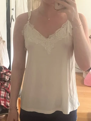 Beige linnetopp med spetsdetaljer - Säljer en ljusbeige linnetopp med tunna axelband och vacker spets längs v-ringningen och upptill på ryggen. Toppen har en avslappnad passform och är perfekt för varma dagar. Superfin att styla med jeans eller kjol!