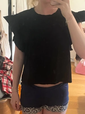 Svart topp med volangärmar från Zara - Säljer en svart topp från Zara med volangdetaljer på axlarna. Toppen har en croppad passform och är tillverkad i mjuk bomull. Perfekt för dig som gillar en enkel men ändå trendig stil.