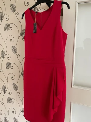 Röd ärmlös klänning med volang - Snygg röd klänning med v-ringning och ärmlös design. Klänningen har en kort längd och en dekorativ volangdetalj på sidan som ger en cool touch. Perfekt för dig som vill sticka ut med en färgstark look.