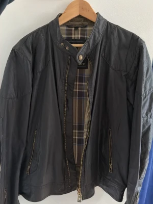 Svart vaxjacka från Belstaff - Snygg svart vax jacka från Belstaff med rutigt innerfoder och dragkedjor framtill. Jackan har ståkrage med knapp och två fickor med dragkedja. Perfekt för dig som vill ha en stilren och klassisk look.