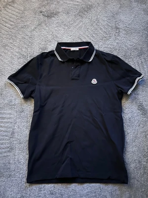 Svart Moncler pikétröja med vit kant - Klassisk svart pikétröja från Moncler med vit kant på krage och ärmslut. Tröjan har korta ärmar, knappslå framtill och Moncler-logga broderad på bröstet. Perfekt för en clean och stilren look.