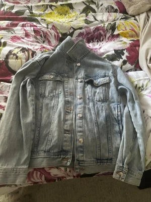 Ljusblå klassisk jeansjacka - Säljer en ljusblå jeansjacka med raka sömmar och silverfärgade knappar. Jackan har två bröstfickor med lock och knapp samt klassisk krage. Perfekt oversized look och tidlös design som funkar till allt.