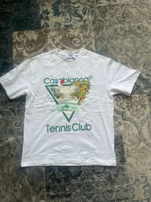 Vit Casablanca Tennis Club t-shirt storlek S - Snygg vit t-shirt från Casablanca med trycket 'Tennis Club' och ett färgglatt motiv framtill. Klassisk passform med korta ärmar och rund hals. Perfekt för dig som gillar sportig och trendig stil. Den har storlek S, är helt ny och aldrig buren