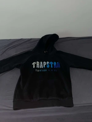 Svart hoodie från Trapstar - Svart hoodie från Trapstar med huva och stor ficka framtill. Trycket på bröstet har Trapstar-loggan i gradient från vitt till blått och texten 'It's a secret' under. Materialet är mjukt och bekvämt, perfekt för en avslappnad streetwear-look.