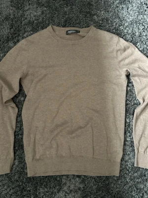 Beige stickad tröja från Dressmann - En stilren beige stickad tröja från Dressmann med rund halsringning och långa ärmar. Tröjan har ribbade muddar vid ärmslut och nederkant, vilket ger en klassisk och enkel look. Perfekt att matcha med jeans eller chinos.