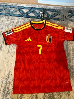 Belgien matchtröja De Bruyne #7 - Snygg Belgien matchtröja från Adidas med De Bruyne och nummer 7 på ryggen. Tröjan är röd med gula detaljer, svarta inslag och ett eldliknande mönster. Korta ärmar, officiella märken på ärmarna och klubbmärke på bröstet. Perfekt för fotbollsfans!