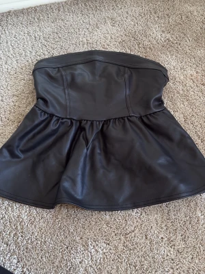 Svart axelbandslös peplum tubtopp - Snygg svart axelbandslös tubtopp i syntetmaterial med peplumskärning. Toppen har dragkedja bak för enkel påklädning och en lätt glansig finish som ger en edgy look. Perfekt att styla med jeans eller kjol för en trendig outfit.