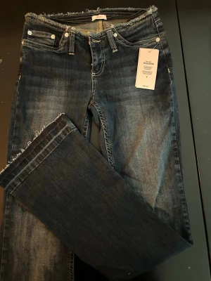 Low Rise/Waist jeans från Nelly - Skitsnygga mörkblå low waist jeans från Nelly med lite bootcut och råa, fransiga kanter i midja och benslut. Klassisk femficksmodell med vita sömmar och knappgylf. Stretchiga. Säljer endast då de inte passade mig, oanvända. 600 nypris