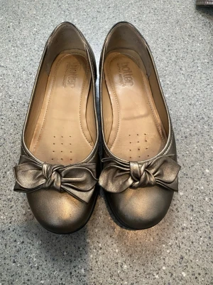 Bronsfärgade ballerinaskor med rosett - Snygga ballerinaskor från Hotter i metallicfärgat material med rund tå och dekorativ rosett framtill. Skorna har en mjuk innersula och svart sula för extra komfort. Perfekta för dig som vill ha en stilren och trendig look. Kolla gärna mina andra annonser❗️
